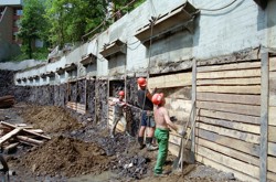 Realizace záporového pažení hluboké stavební jámy. Postupné osazování dřevěných pažin mezi jednotlivé zápory za současného snižování výkopů na další kotevní úroveň.